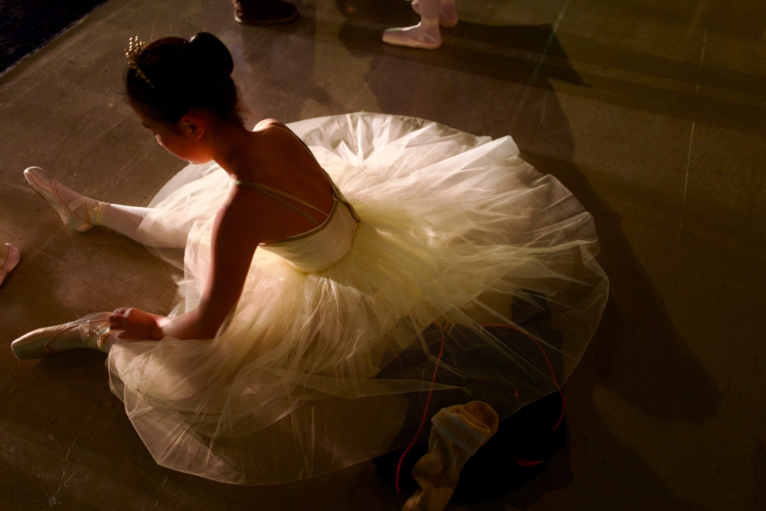 girl dressed in ballerina costume sitting on floor facing away from camera