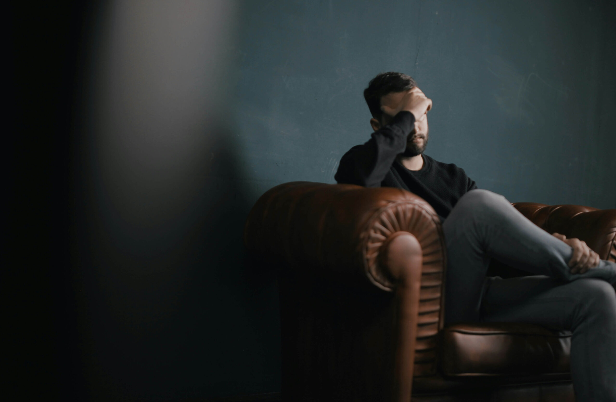 adult male sitting on couch with legs crossed and hand on forehead.