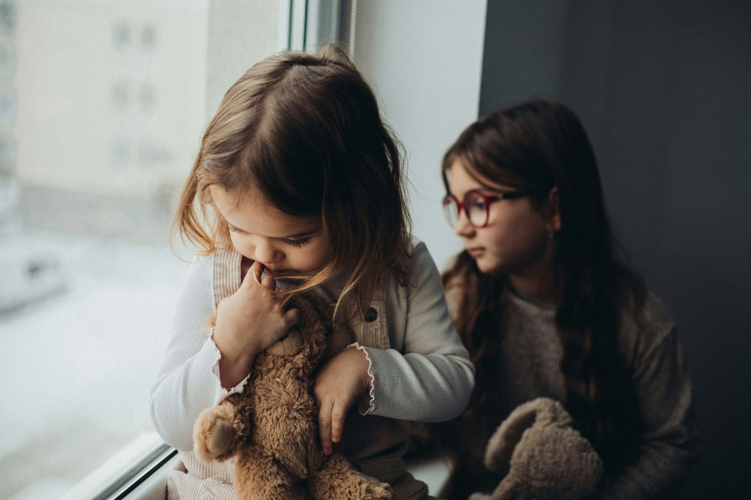 two young kids looking out window hugging stuffed animal